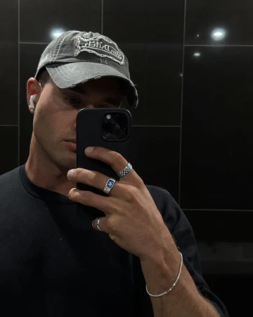 Selfie of a young adult man wearing a baseball cap and rings, holding a phone in an indoor setting with dark tiled background.