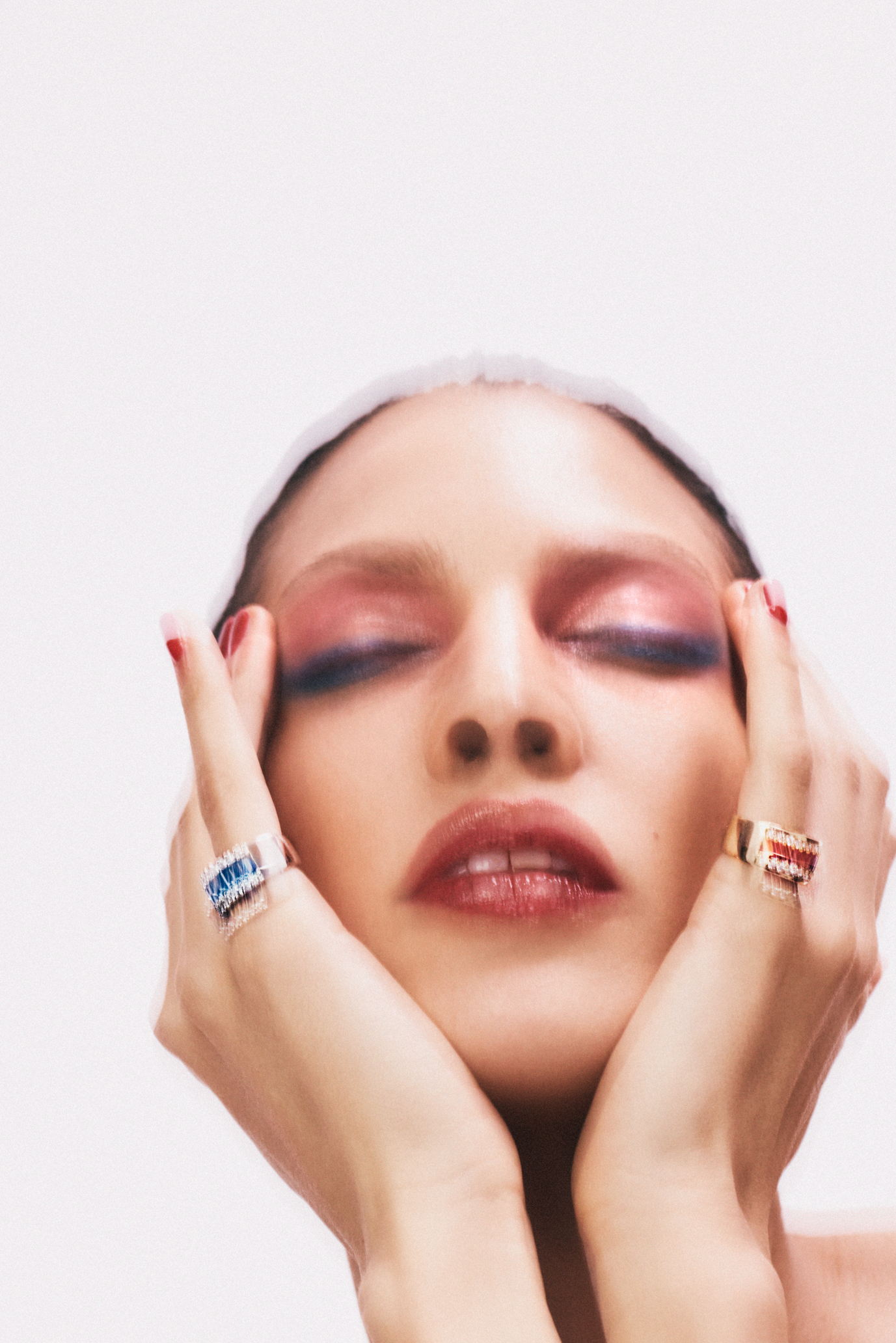 Close-up studio shot of a fair-skinned young adult woman with closed eyes and colorful eyeshadow, wearing rings and a white head covering.