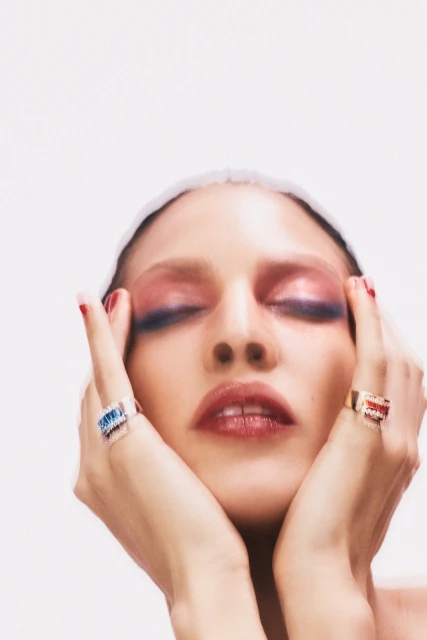 Close-up studio shot of a fair-skinned young adult woman with closed eyes and colorful eyeshadow, wearing rings and a white head covering.
