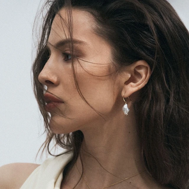 Close-up shot of a young adult woman with dark hair wearing a pearl earring and necklace, on a neutral background.