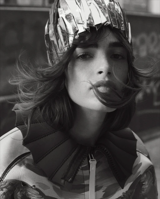 Close-up black and white portrait of a woman wearing a metallic headpiece and jacket with large collar, captured at eye-level.