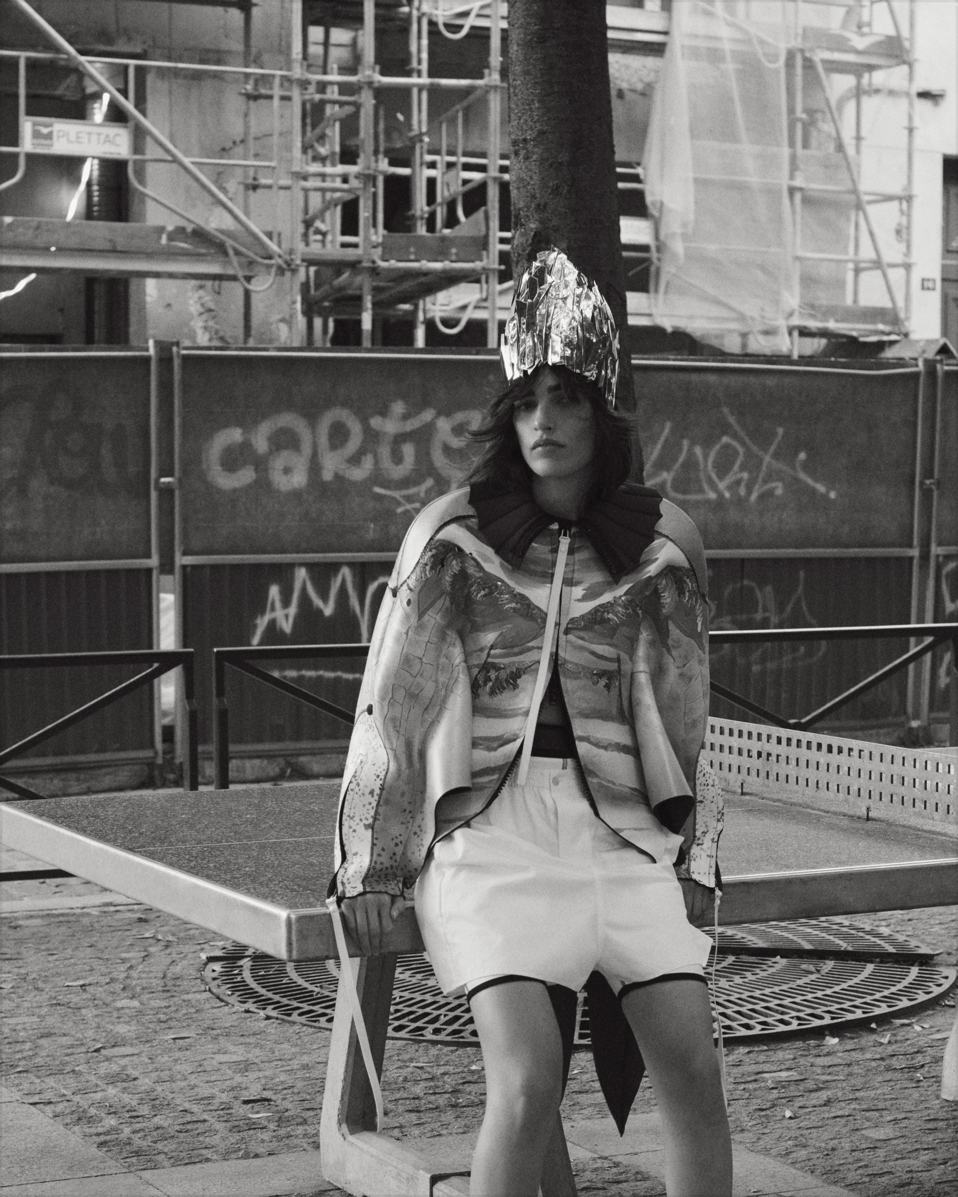 A young woman with dark hair poses outdoors on a stone bench, wearing a shiny headdress, patterned top, and white shorts in black and white.
