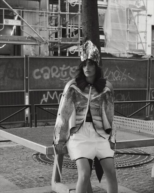A young woman with dark hair poses outdoors on a stone bench, wearing a shiny headdress, patterned top, and white shorts in black and white.