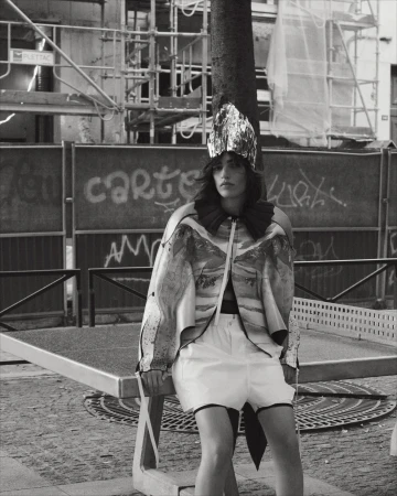 A young woman with dark hair poses outdoors on a stone bench, wearing a shiny headdress, patterned top, and white shorts in black and white.