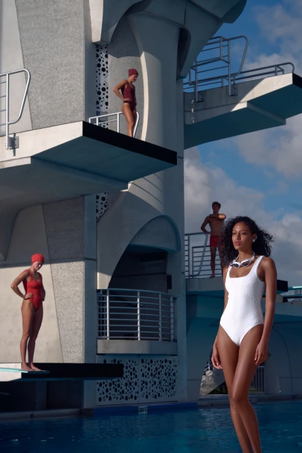 Four people in swimsuits near a diving platform and swimming pool, wearing red and white swimsuits with sky and clouds background.