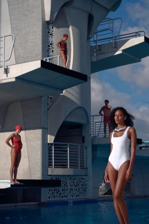 Four people in swimsuits near a diving platform and swimming pool, wearing red and white swimsuits with sky and clouds background.