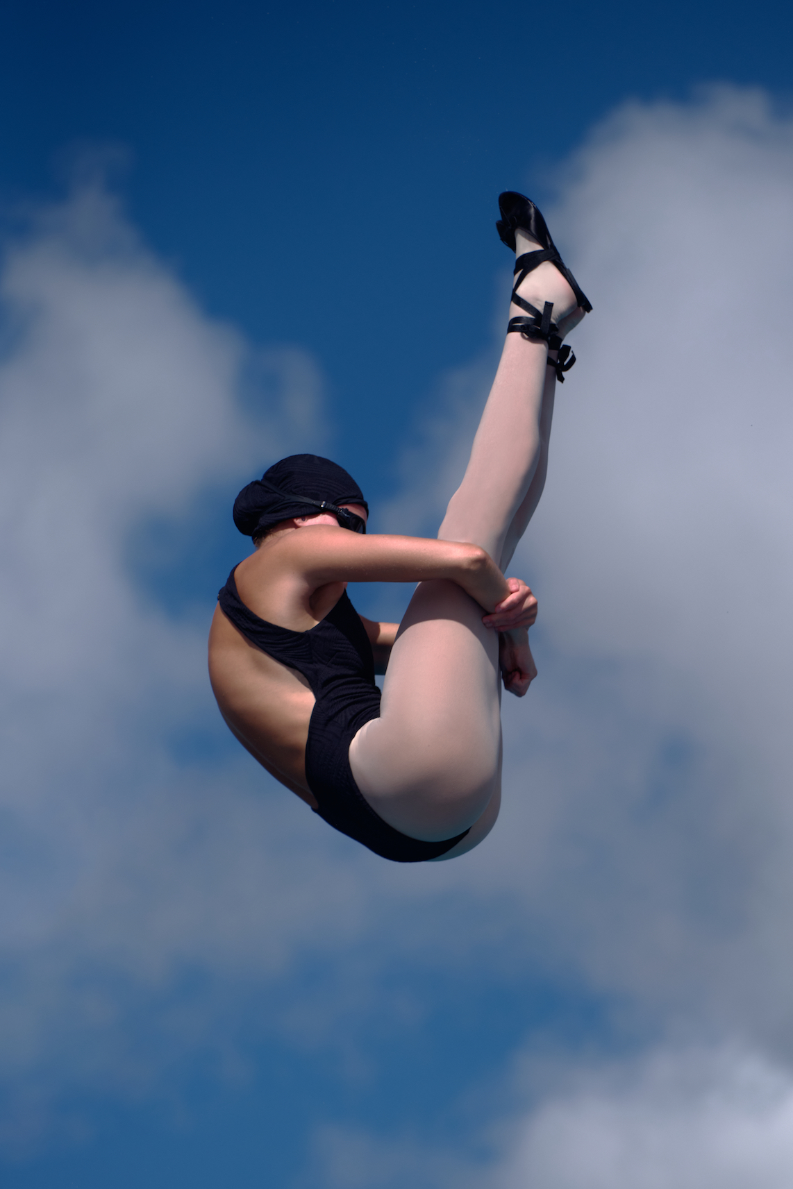 Female athlete performs ballet pose against a vibrant blue sky with clouds. She wears black leotard, light tights, and black ballet shoes.