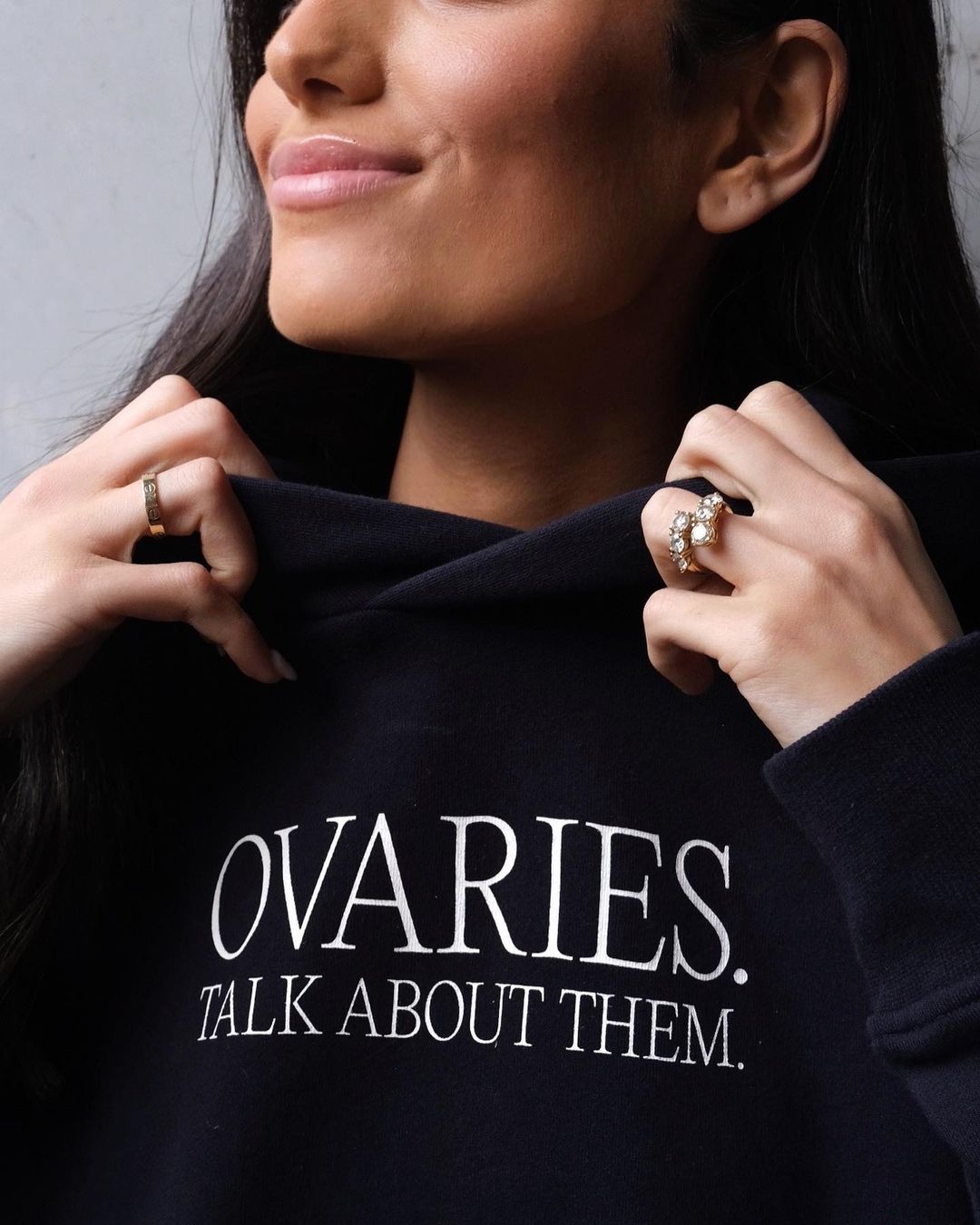 Young adult female wearing a black hoodie with white text and jewelry posing indoors with a neutral background.
