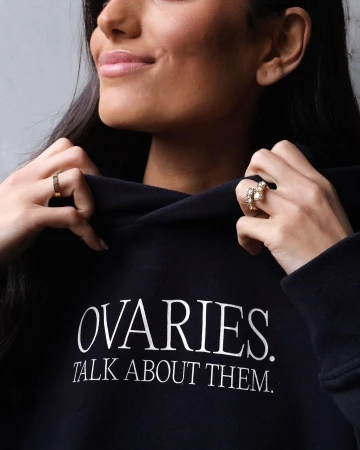 Young adult female wearing a black hoodie with white text and jewelry posing indoors with a neutral background.