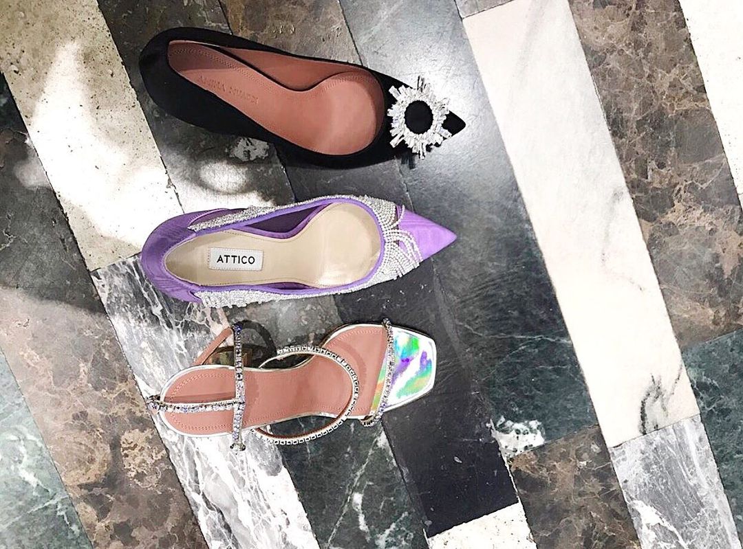 Three luxury designer shoes are arranged on a marble floor in a close-up shot. Black, lavender, and iridescent heels are visible.