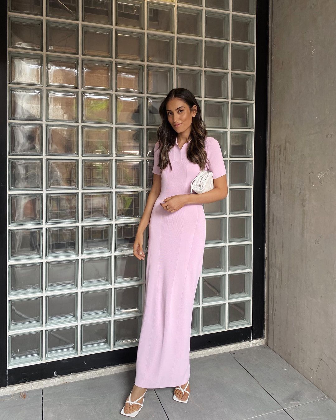 Woman in light pink knit dress and white heels standing in front of wall with glass block in outdoor setting with natural light.