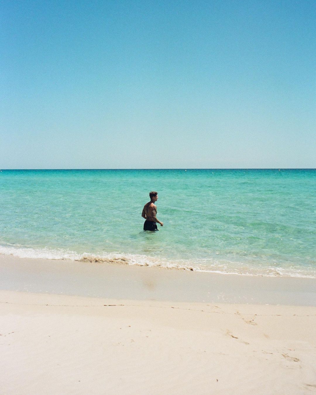 A shirtless man in dark swim shorts wading in shallow turquoise ocean water on a sandy beach.