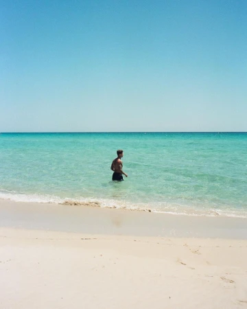 A shirtless man in dark swim shorts wading in shallow turquoise ocean water on a sandy beach.