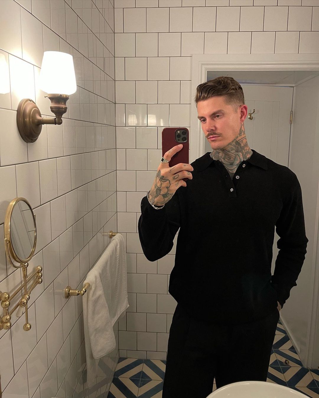 A man with tattoos in a black outfit poses in a bathroom with white tiled walls and a patterned tile floor while taking a selfie.
