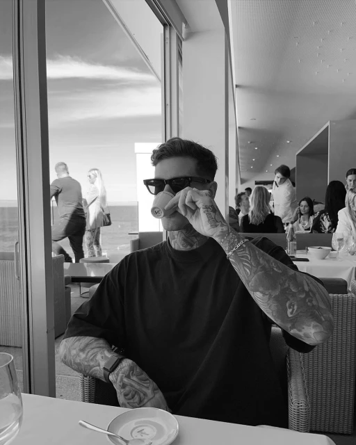 Man with tattoos indoors at restaurant with other patrons, drinking from a small cup, monochromatic, near a large glass window.