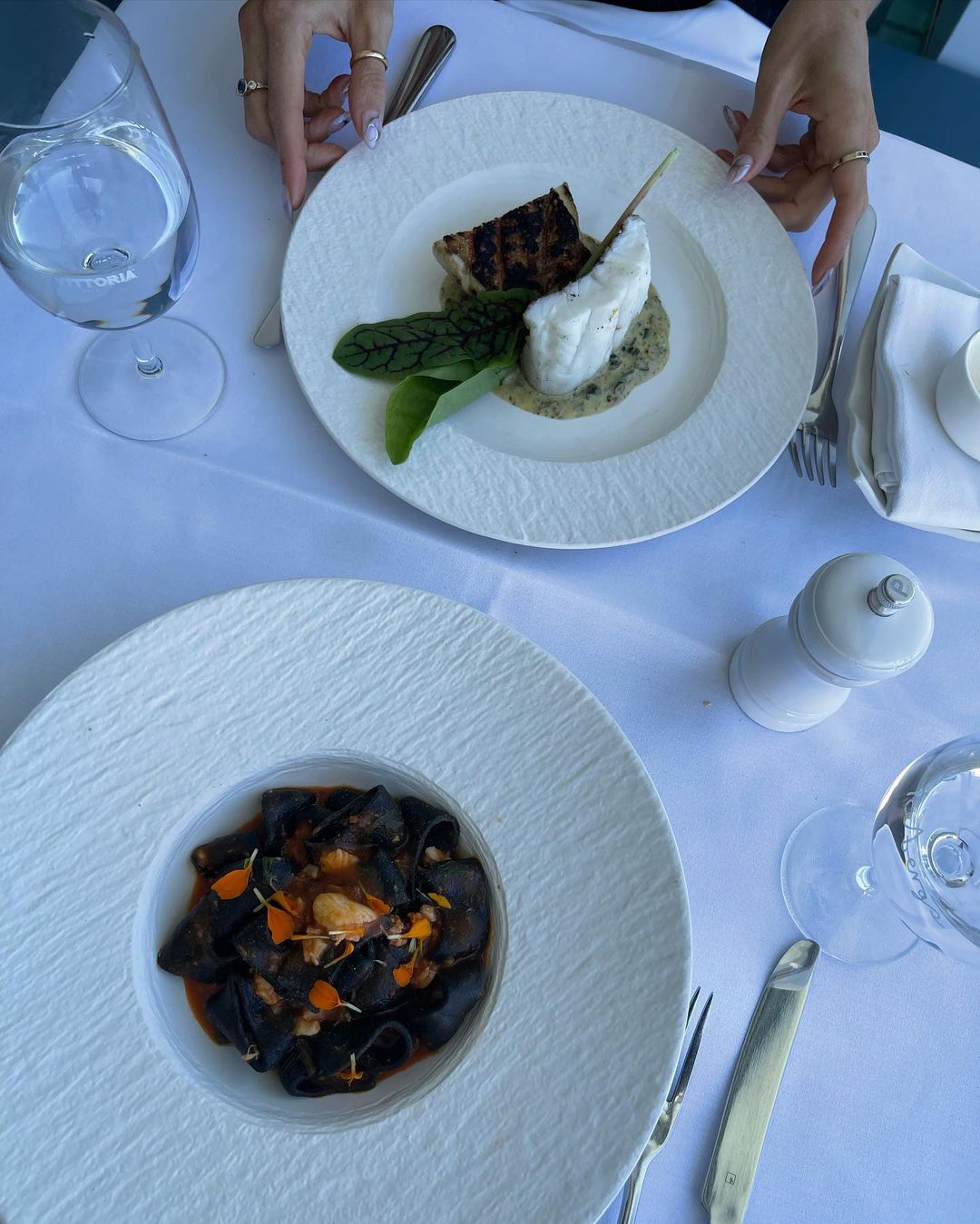 Overhead shot of two plates with fish and dark pasta on a table at a restaurant, with silverware, water glasses, and salt shaker.