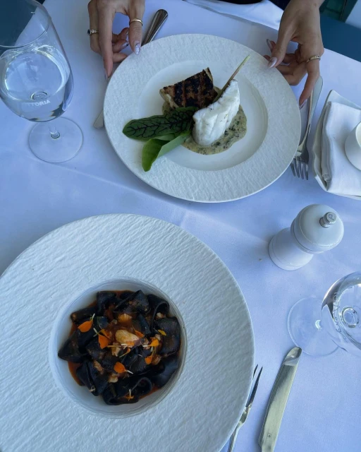 Overhead shot of two plates with fish and dark pasta on a table at a restaurant, with silverware, water glasses, and salt shaker.