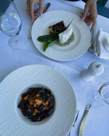 Overhead shot of two plates with fish and dark pasta on a table at a restaurant, with silverware, water glasses, and salt shaker.