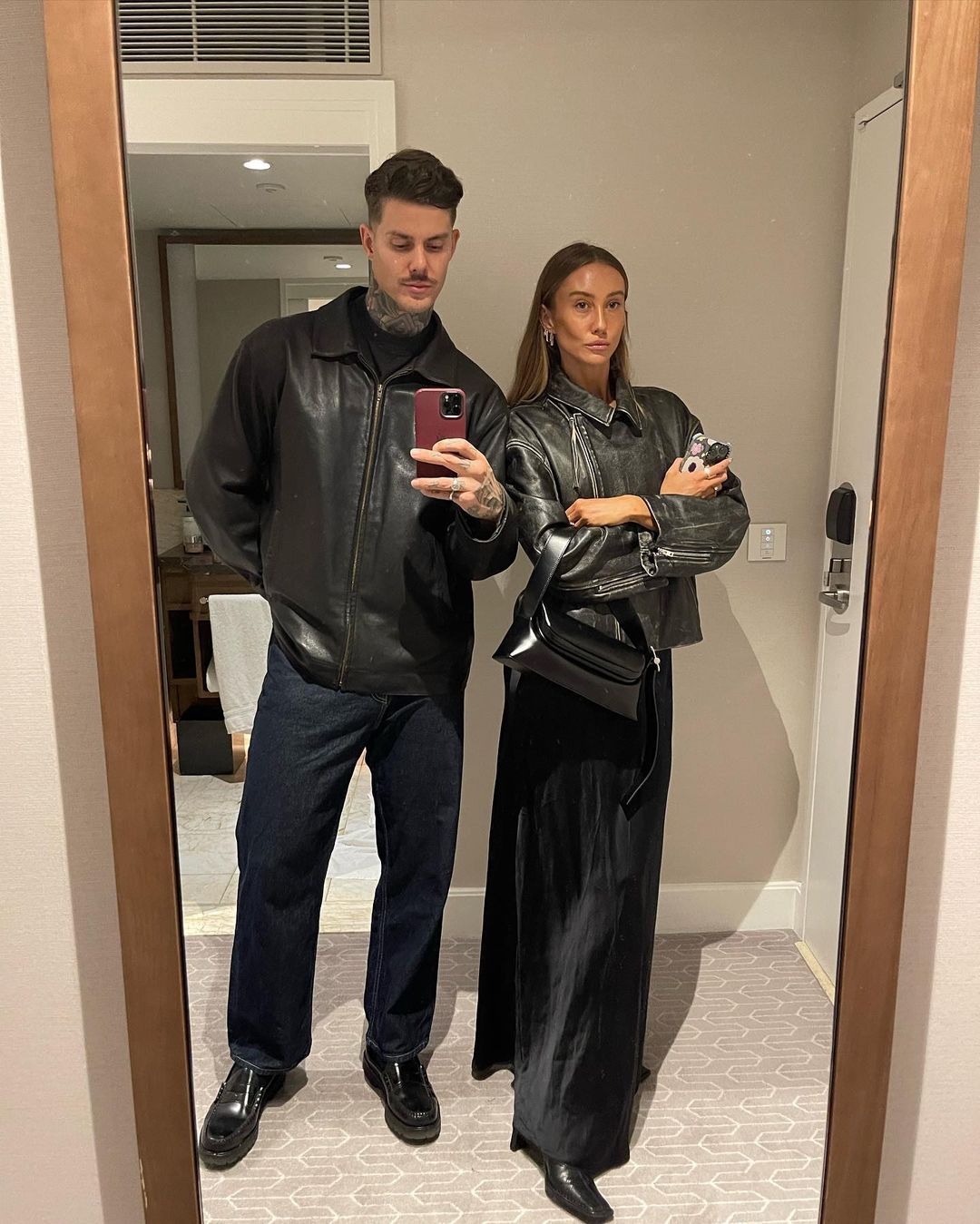 Indoor portrait of a fashionable couple wearing leather jackets and dark trousers standing in front of a mirror, looking at the camera.