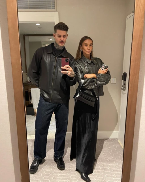 Indoor portrait of a fashionable couple wearing leather jackets and dark trousers standing in front of a mirror, looking at the camera.