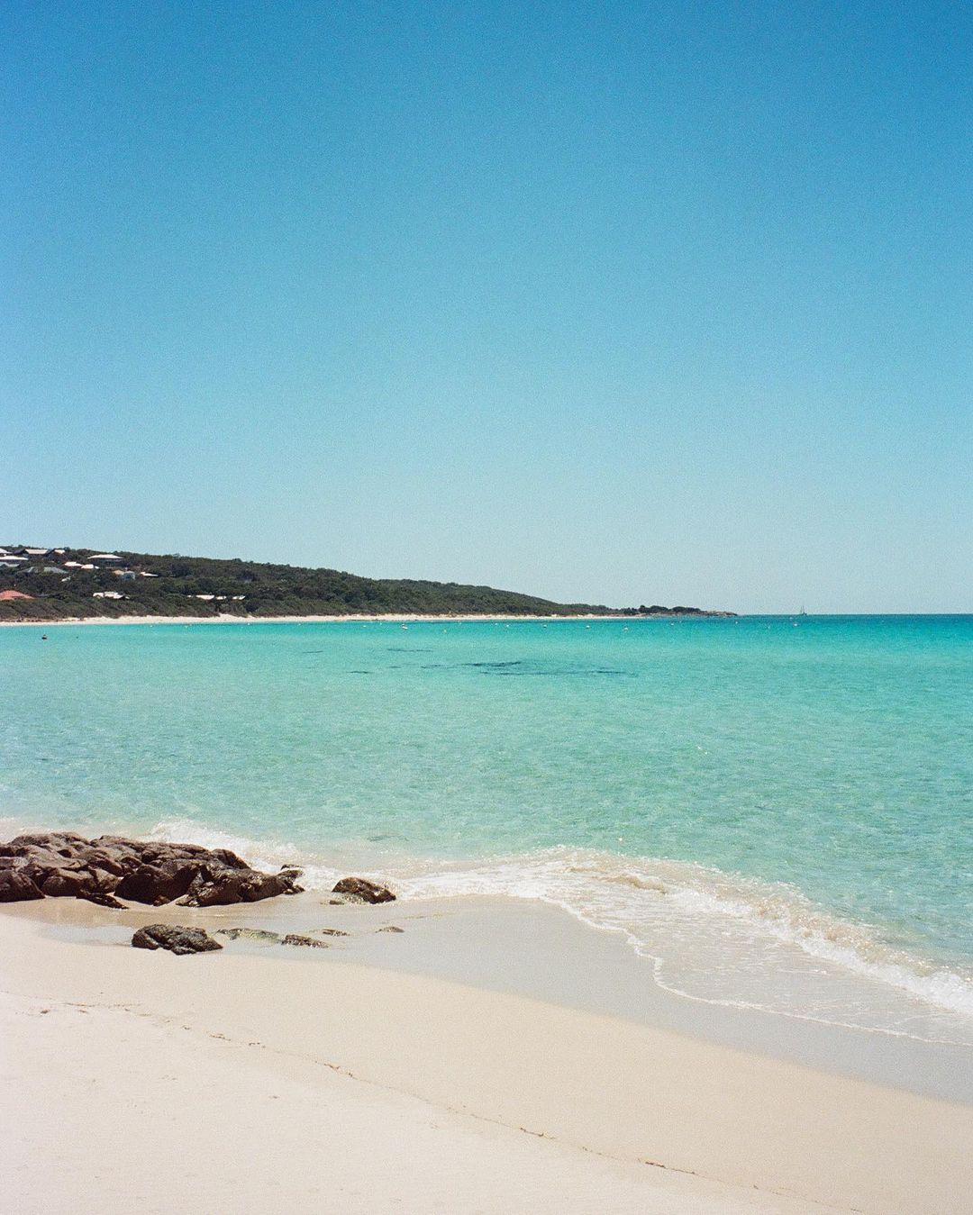 Sunny coastal view of a sandy beach with turquoise ocean water, gentle breaking waves, and dark rocks under a clear blue sky