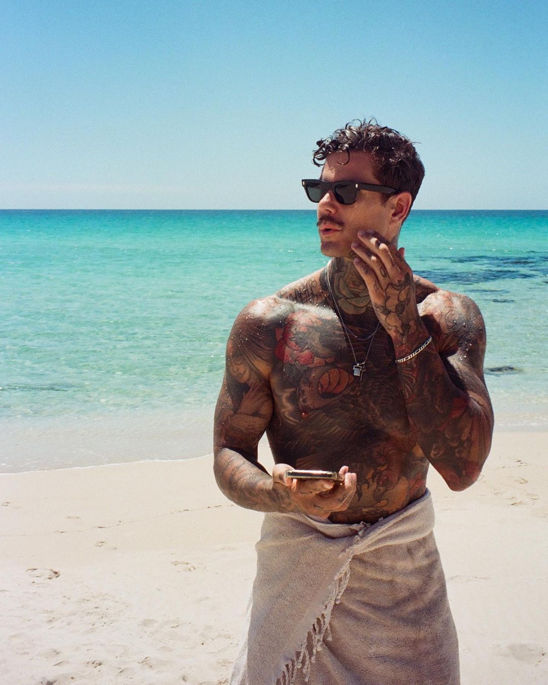 Tattooed man on beach wearing sunglasses and wrap. Tropical ocean scene with clear turquoise water and white sand under a sunny blue sky.