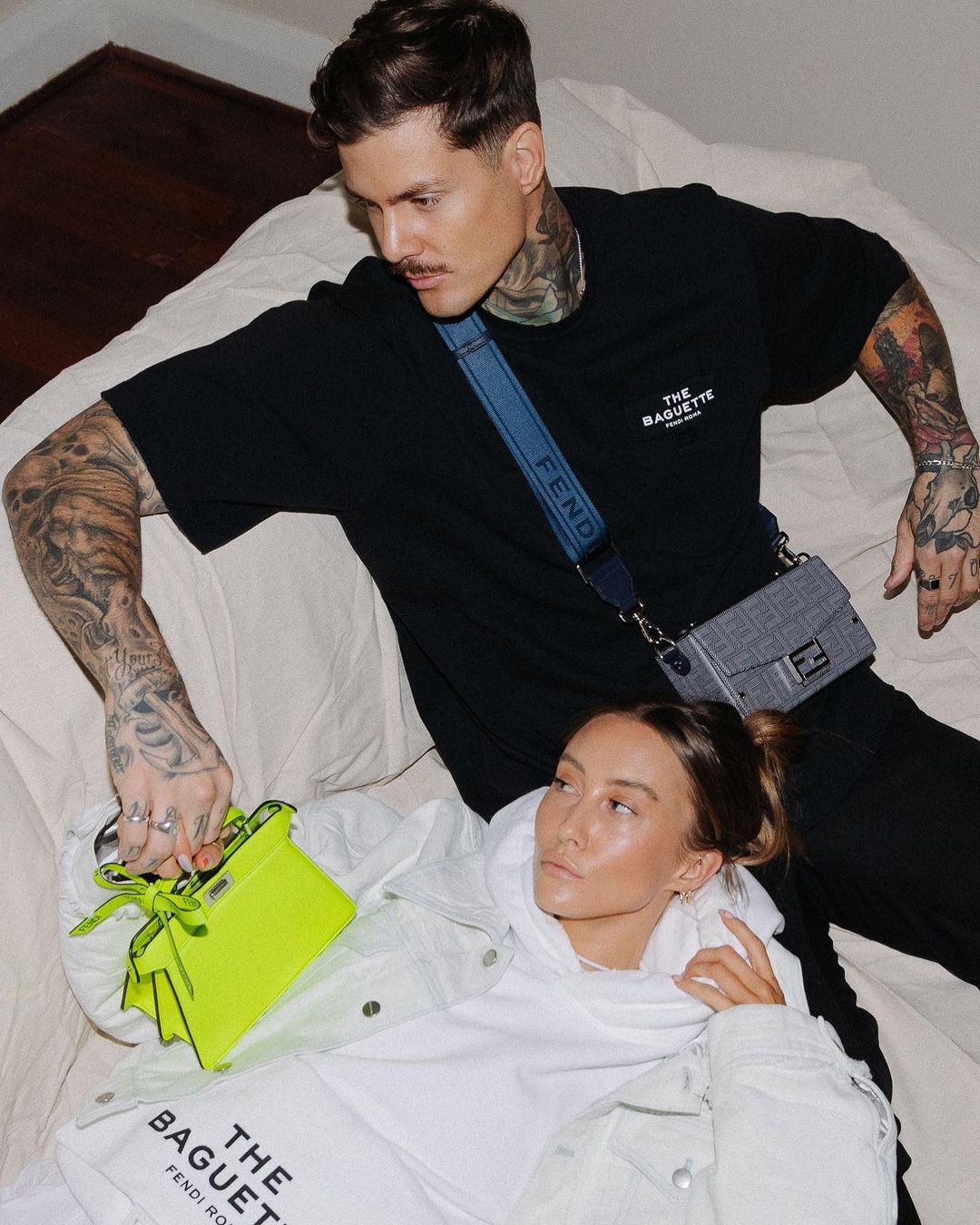 Two fashion models pose indoors on a bed, showcasing Fendi apparel and accessories, including a black t-shirt, white sweatshirt and neon green purse.