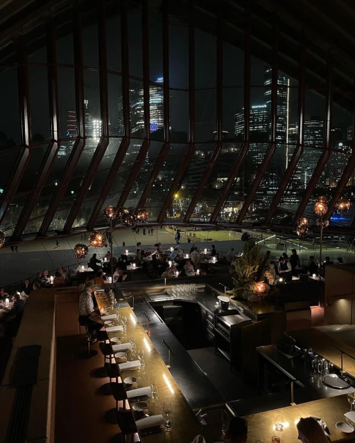 Indoor dining at a restaurant with a bar and a night cityscape seen through large windows, creating an elegant and inviting atmosphere.