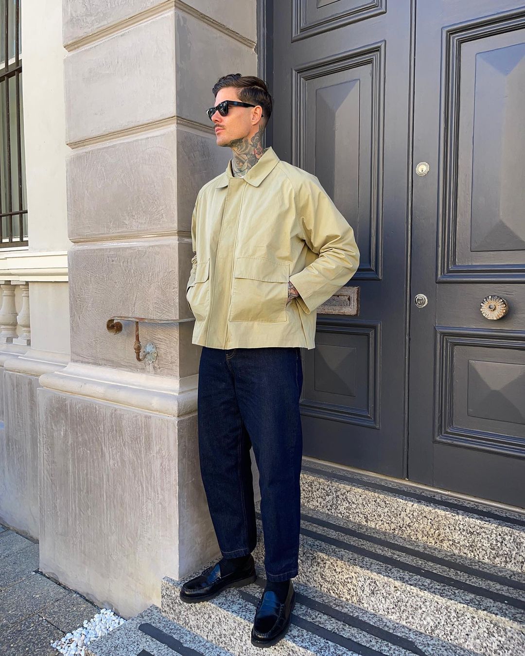 Adult male in jacket and jeans standing on stone steps with dark door and stone building in background in daylight.