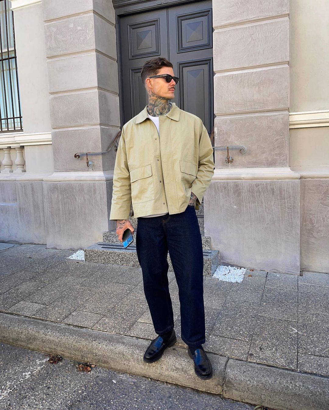Adult male poses outdoors wearing sunglasses, a jacket, pants, and dress shoes; he holds a phone in his left hand and stands against a building.