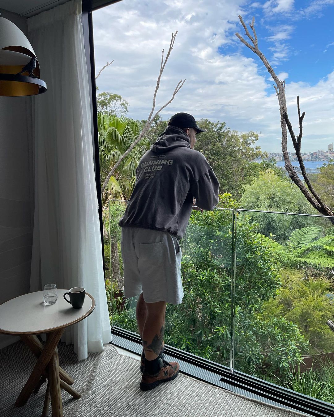 An adult male stands looking out of a glass balcony at a green landscape. A table and coffee cup are visible inside the room.