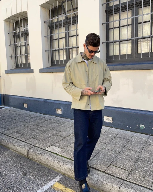 Man in a beige jacket using a phone outdoors in an urban environment, with dark jeans and black shoes, in front of a white building.