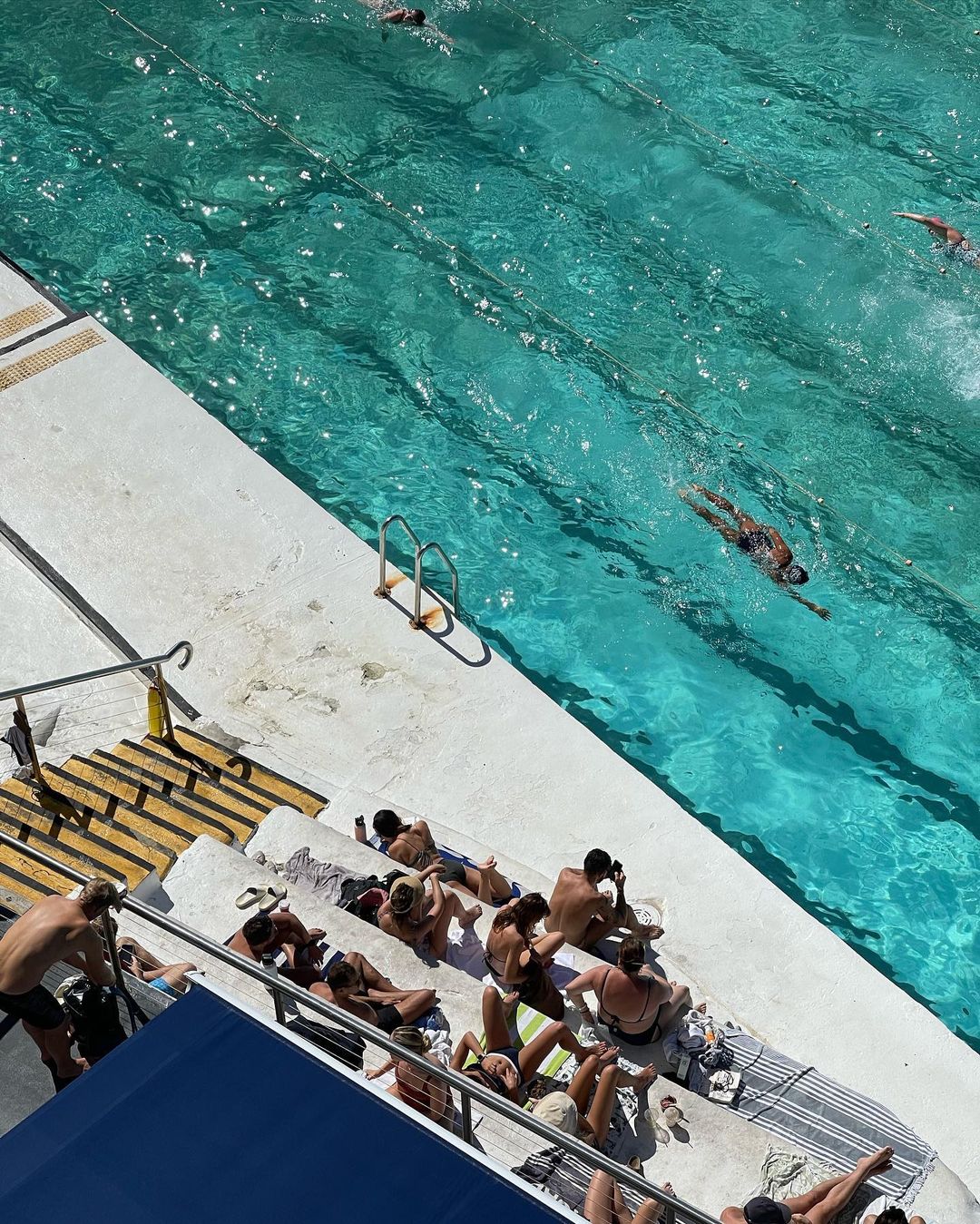 Overhead shot of an outdoor swimming pool with swimmers and people relaxing on the concrete steps, capturing a summer day.