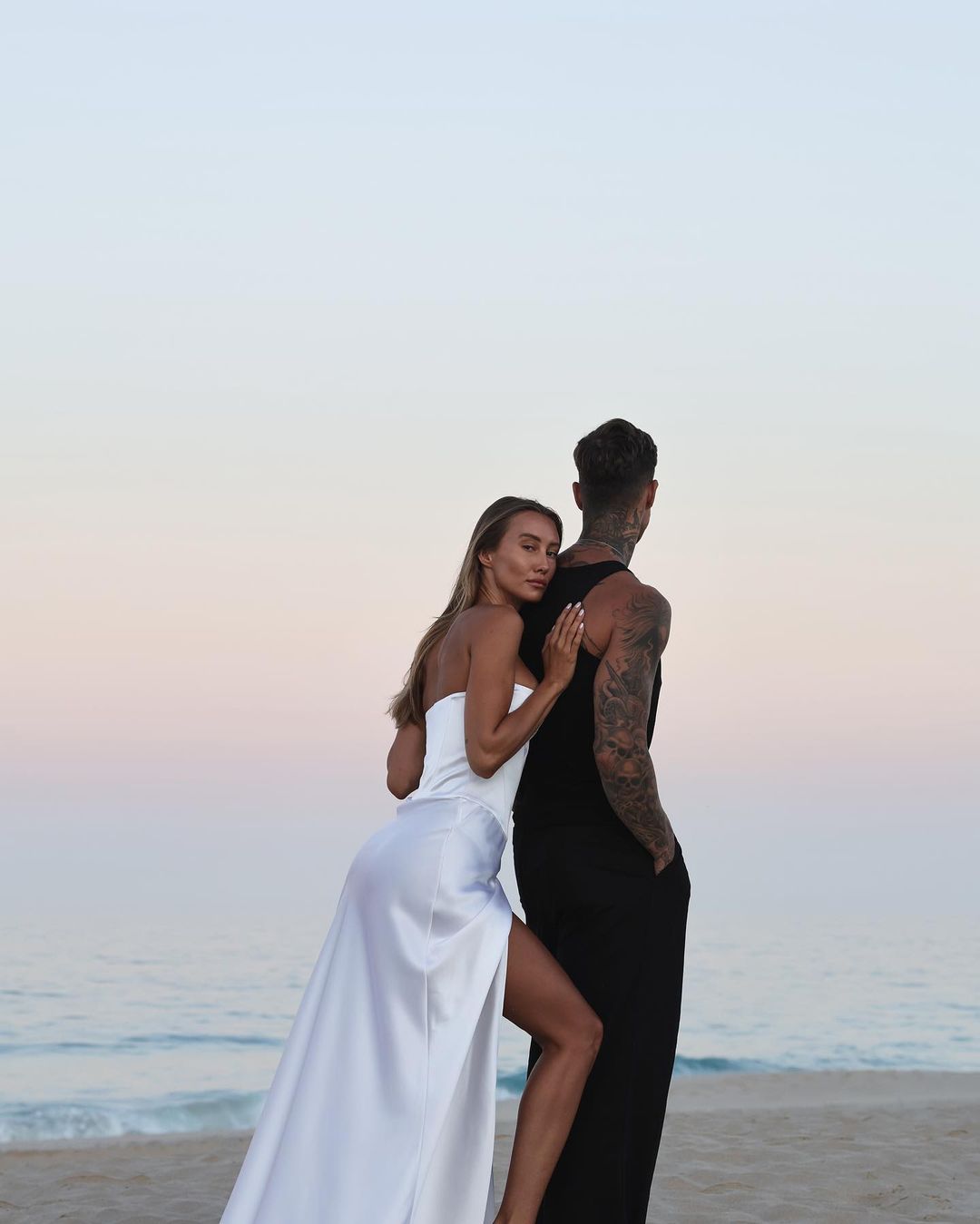Romantic couple on a beach at dusk: a woman in a white dress rests against a man in a black tank dress, ocean and sky backdrop.