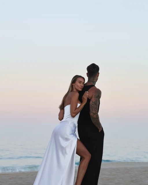 Romantic couple on a beach at dusk: a woman in a white dress rests against a man in a black tank dress, ocean and sky backdrop.