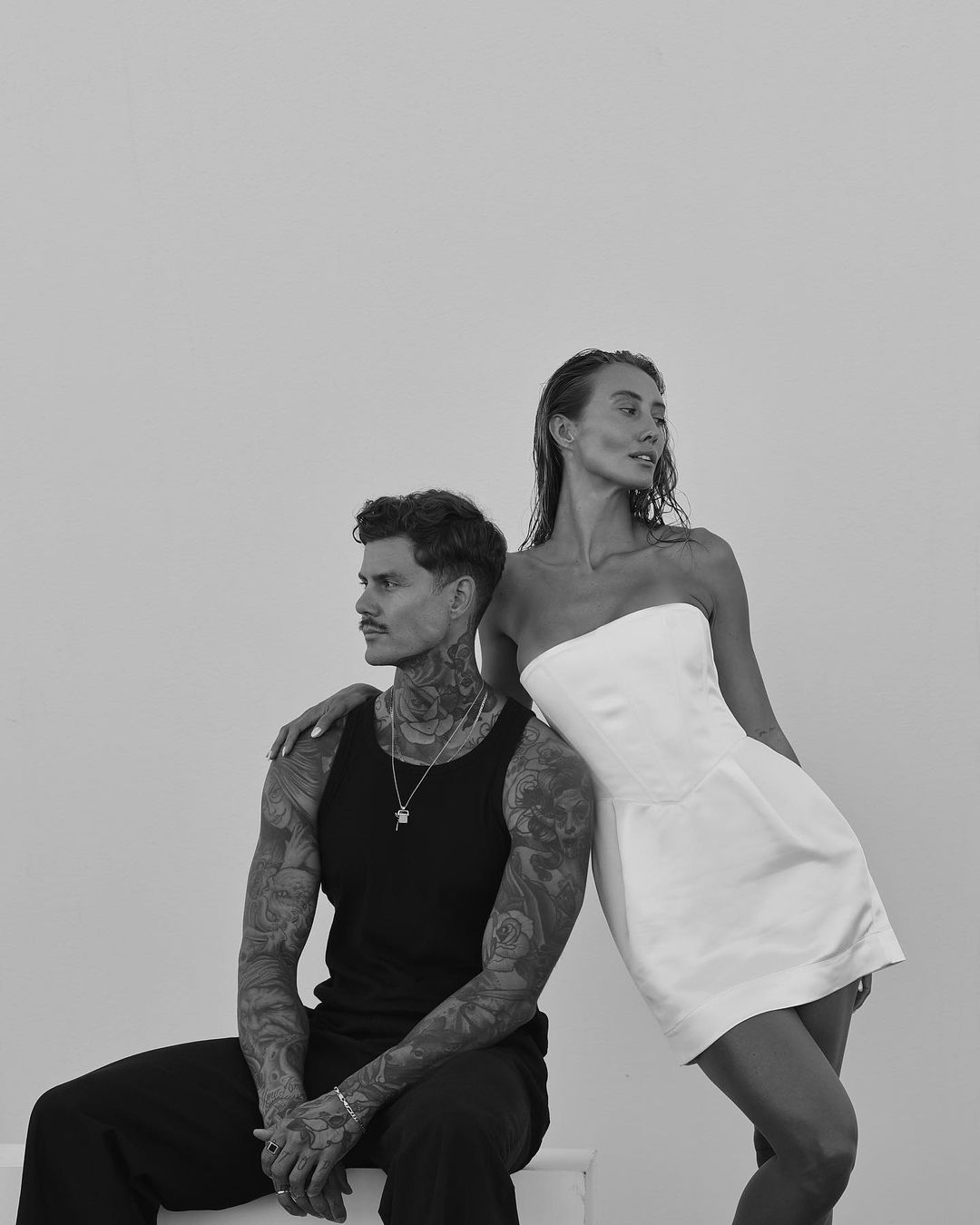 Monochrome portrait of a tattooed man seated in a dark tank top next to a standing woman in a white strapless dress
