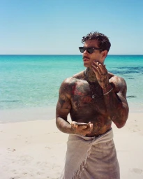 Tattooed man wearing sunglasses and a towel on a sandy beach holding a smartphone by the ocean