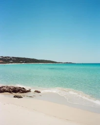 Sunny coastal view of a sandy beach with turquoise ocean water, gentle breaking waves, and dark rocks under a clear blue sky