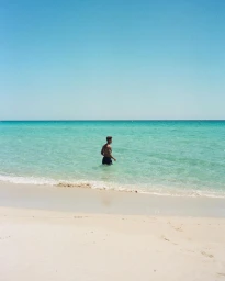 A shirtless man in dark swim shorts wading in shallow turquoise ocean water on a sandy beach.