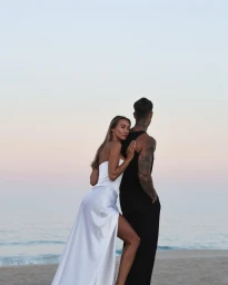 Couple standing on a sandy beach at sunset, a woman in a white satin dress leaning on a man in black attire.