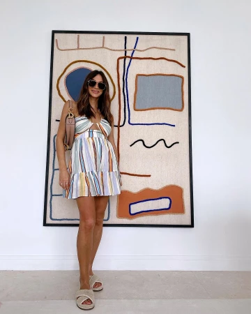 Woman wearing sunglasses and a striped dress standing in front of a large abstract art piece on a white wall