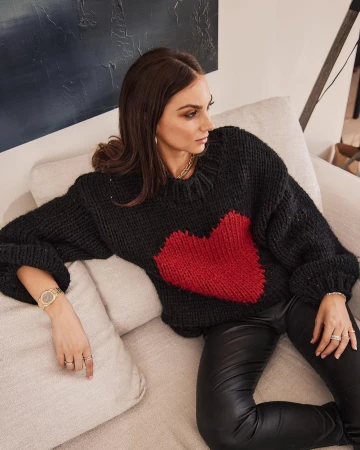 Woman in a black chunky knit sweater with a red heart motif seated on a light beige sofa indoors