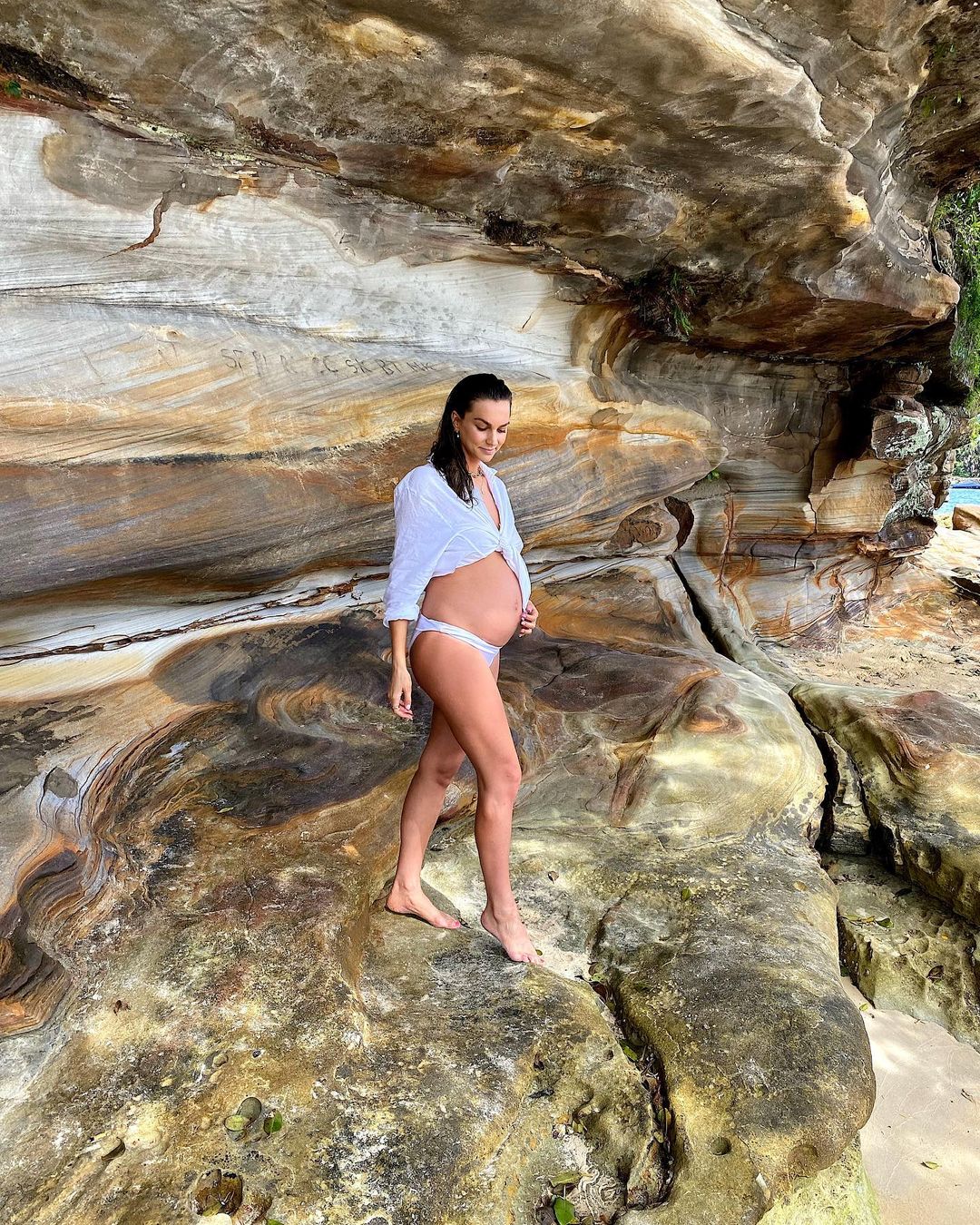 Pregnant woman in a white shirt and swimsuit bottoms standing barefoot on rocky terrain near a beach with visible ocean water.