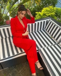 Woman wearing a bright red suit seated on a black and white striped outdoor sofa with palm leaves in the background.