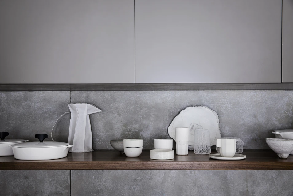 Still life of white tableware including dishes, cups, bowls, and a pitcher, arranged on a wooden shelf against a gray backsplash and cabinets.