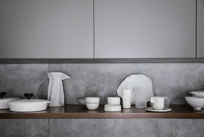 Still life of white tableware including dishes, cups, bowls, and a pitcher, arranged on a wooden shelf against a gray backsplash and cabinets.