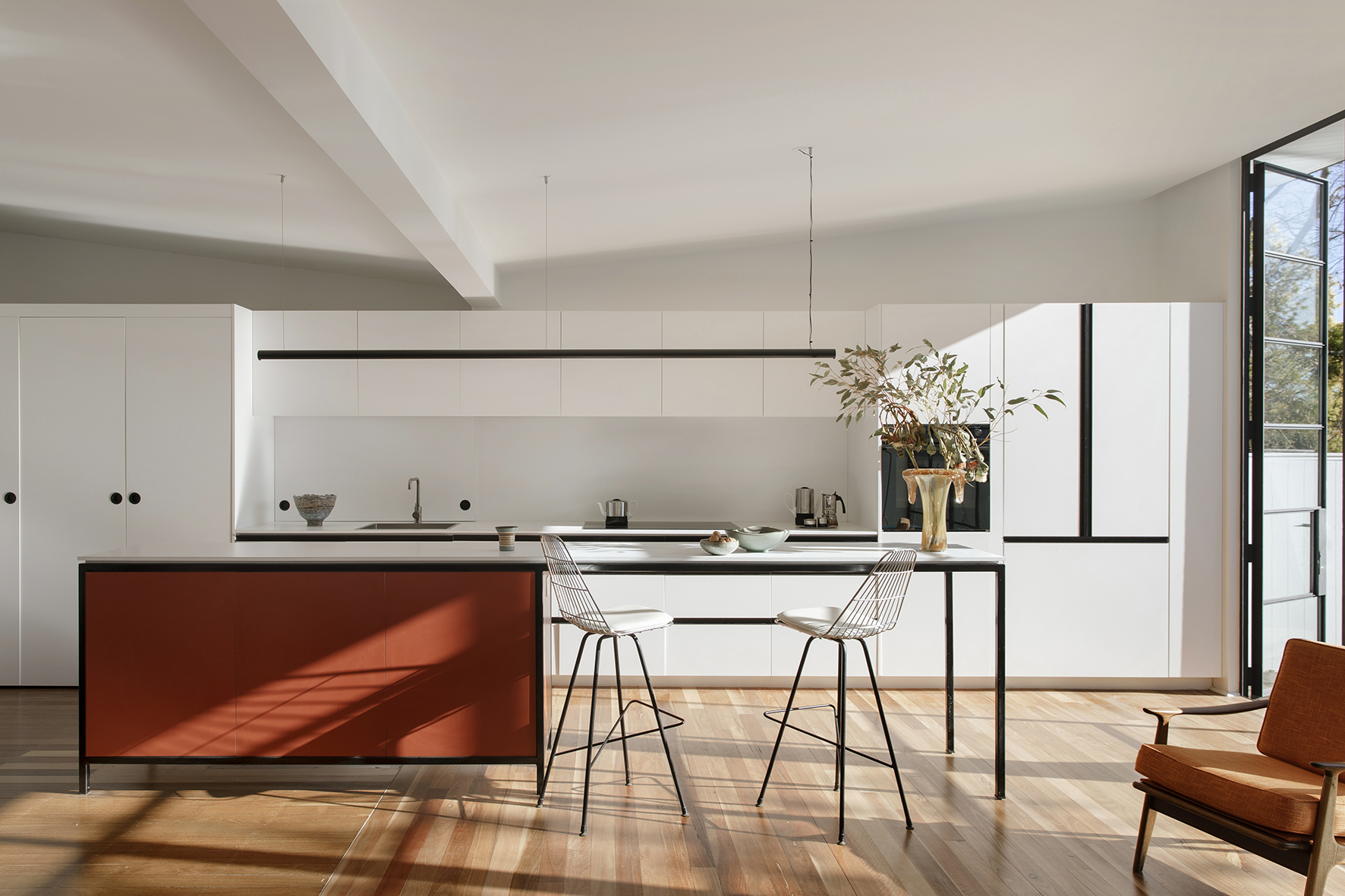 Bright, modern kitchen interior with white cabinets, an orange kitchen island, two stools, hardwood floor, and natural light from a window.
