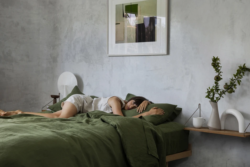 Adult female lying on green bed with green pillows in minimalist home interior. Abstract art hangs above bed with side table.