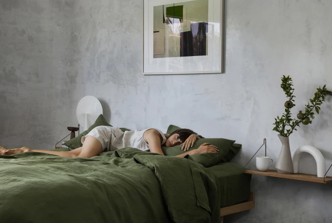 Adult female lying on green bed with green pillows in minimalist home interior. Abstract art hangs above bed with side table.