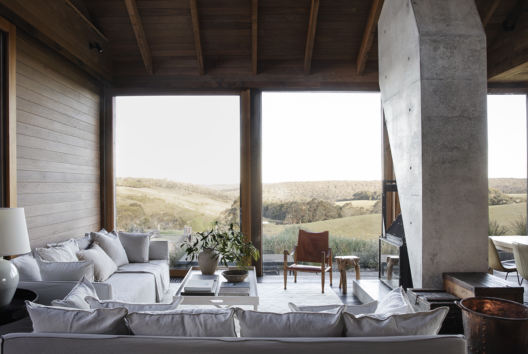 Contemporary living room interior features neutral sofas, a concrete fireplace, a leather chair, and a landscape view through large windows.